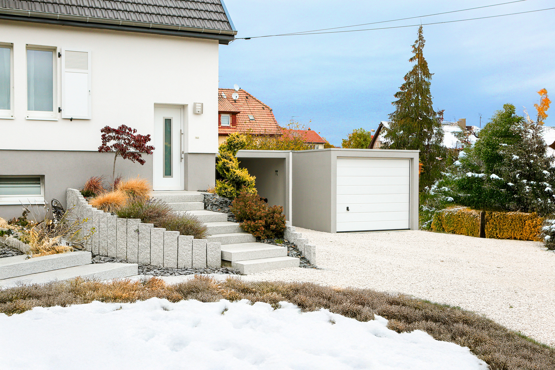 Moderne weiße Einzelgarage mit grauem Vordach, Granitblockstufen und winterlicher Gartengestaltung mit Gräsern
