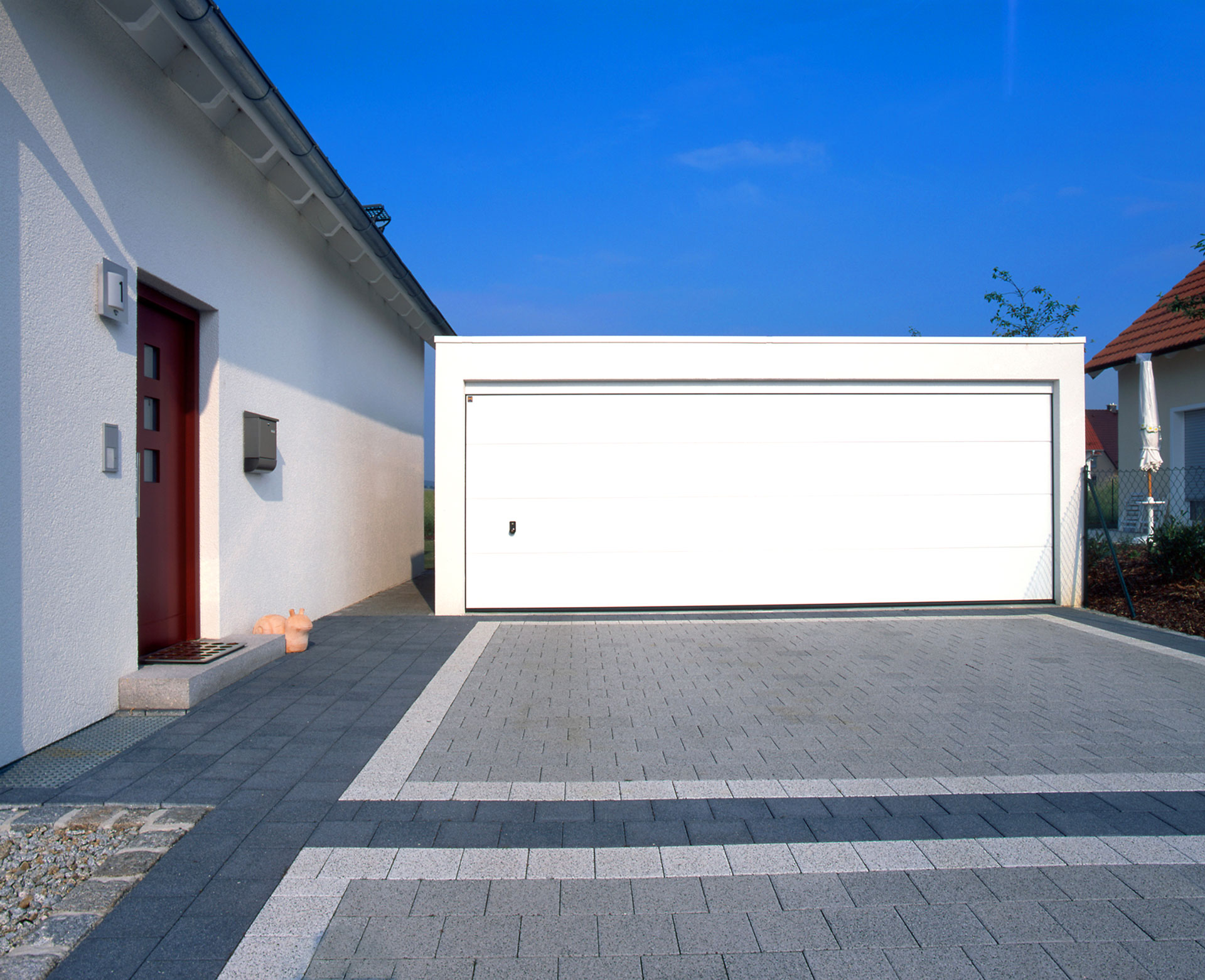 Moderne weiße Großraumgarage mit Sektionaltor und Dachranddesign neben Einfamilienhaus – Fertiggarage aus Beton mit klarer Linienführung