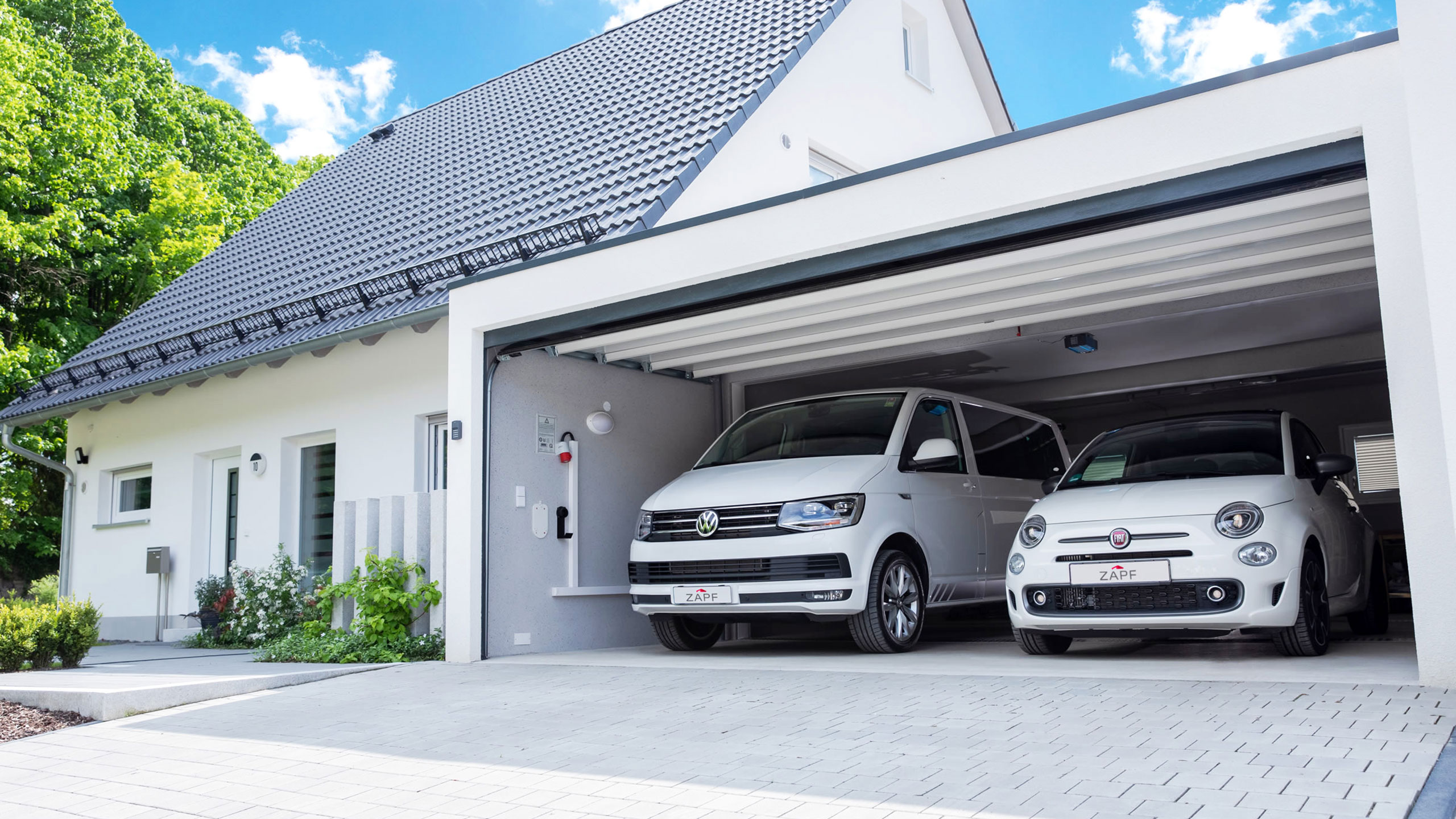 Ein weißer Kleinbus und ein weißes Auto in einer Breitgarage neben einem Einfamilienhaus.