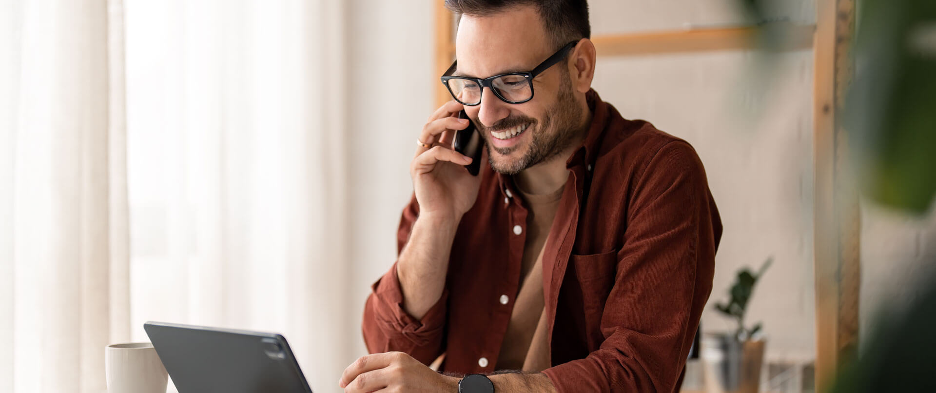 Mann mit Brille telefoniert lächelnd im Homeoffice, sitzt vor Laptop mit Kaffeetasse auf dem Tisch.
