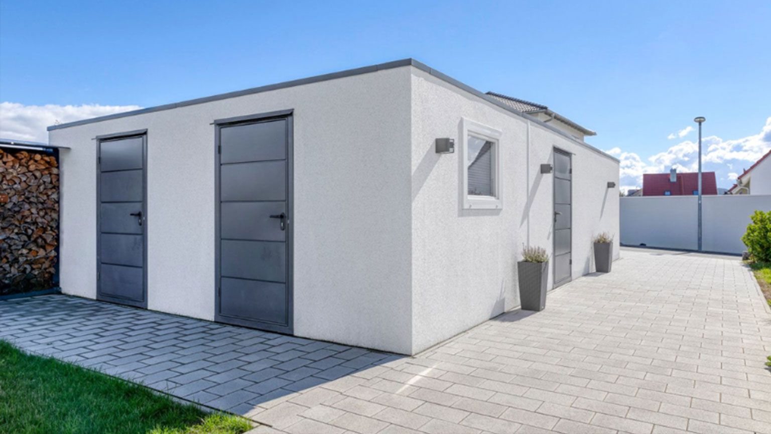 Weiße Garage mit drei modernen Stahltüren, daneben gestapeltes Kaminholz – funktionaler Garagenanbau bei sonnigem Wetter.