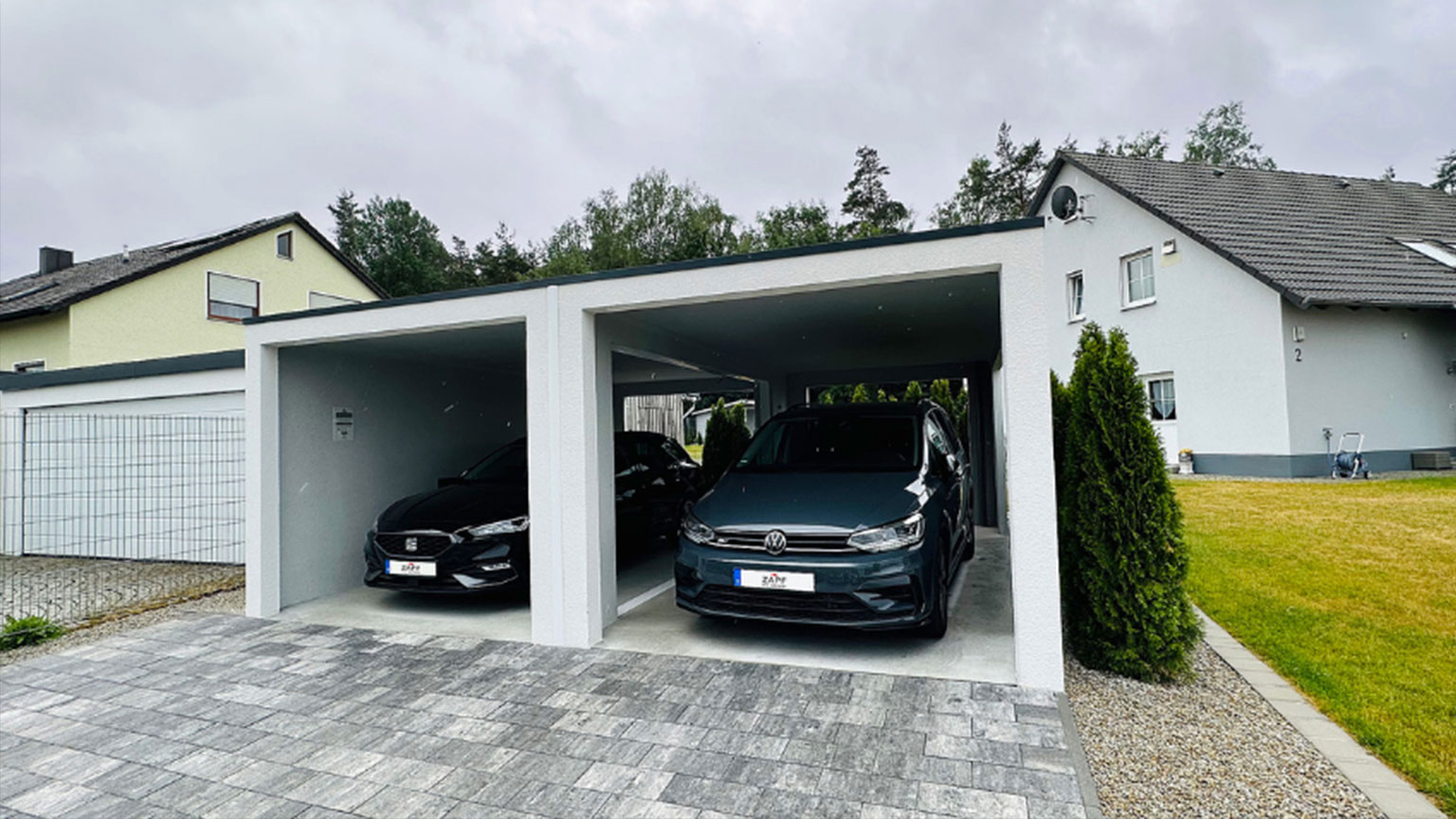 Doppelcarport mit zwei geparkten Fahrzeugen vor einem modernen Einfamilienhaus an einem bewölkten Tag.