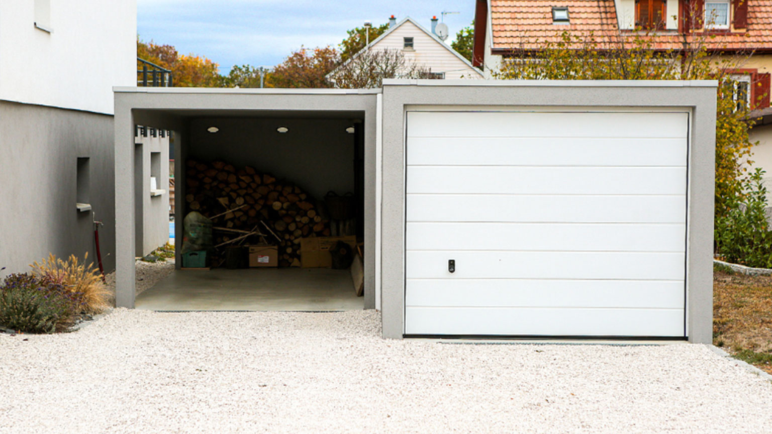Einzelcarport mit integriertem Holzlagerplatz und geschlossener Garage auf hellem Kiesboden.
