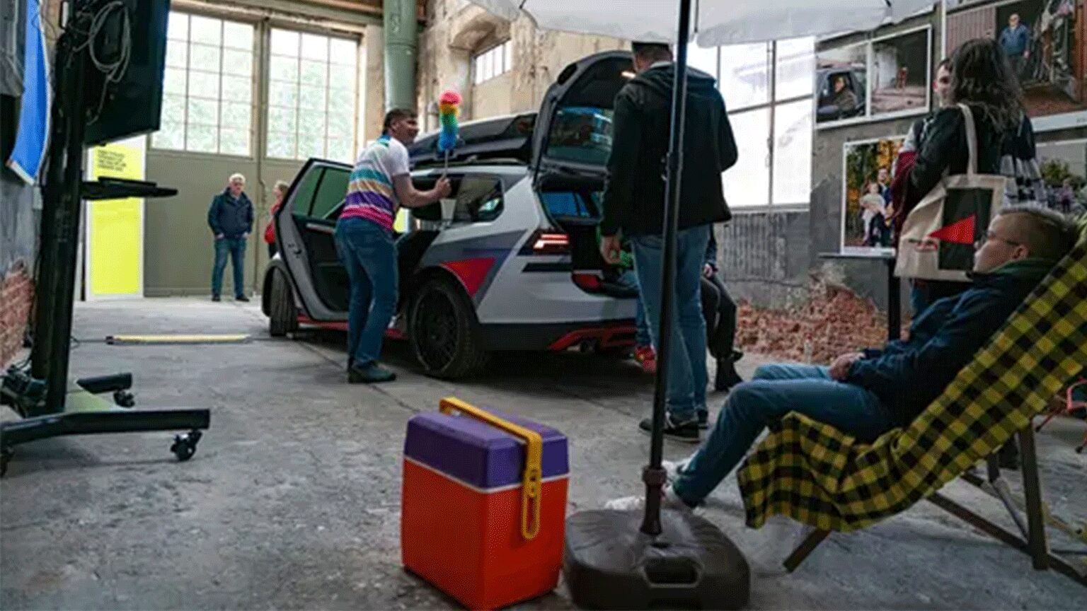 Innenaufnahme eines Fotostudios mit einem Auto als zentrales Motiv, mehreren Personen bei der Arbeit und einem entspannten Zuschauer im Liegestuhl.