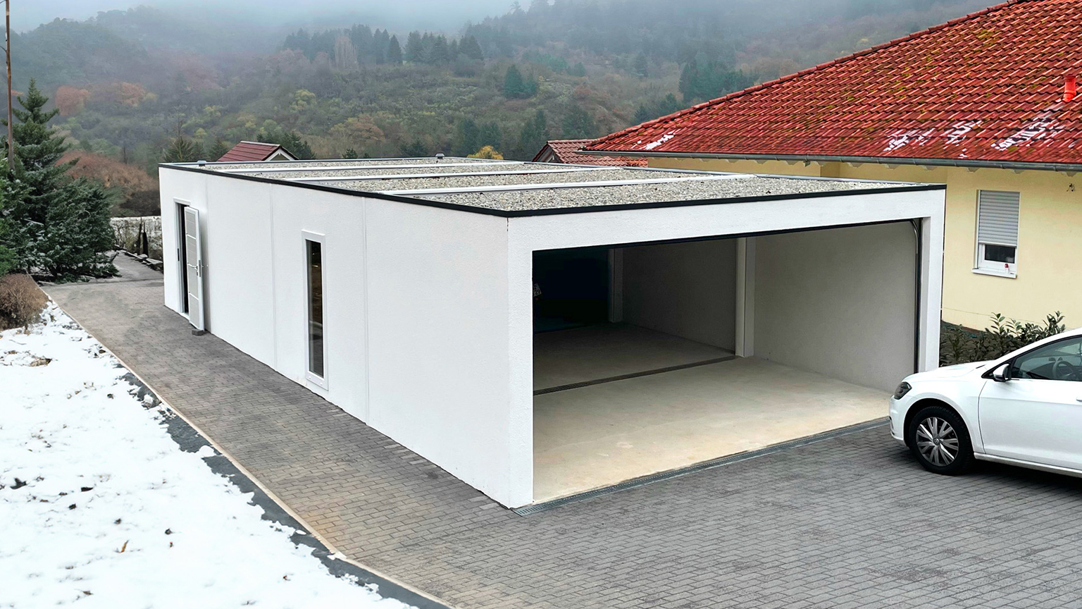 Freistehende moderne Garage mit Flachdach im Winter, teils verschneite Umgebung und geöffnete Toröffnung.