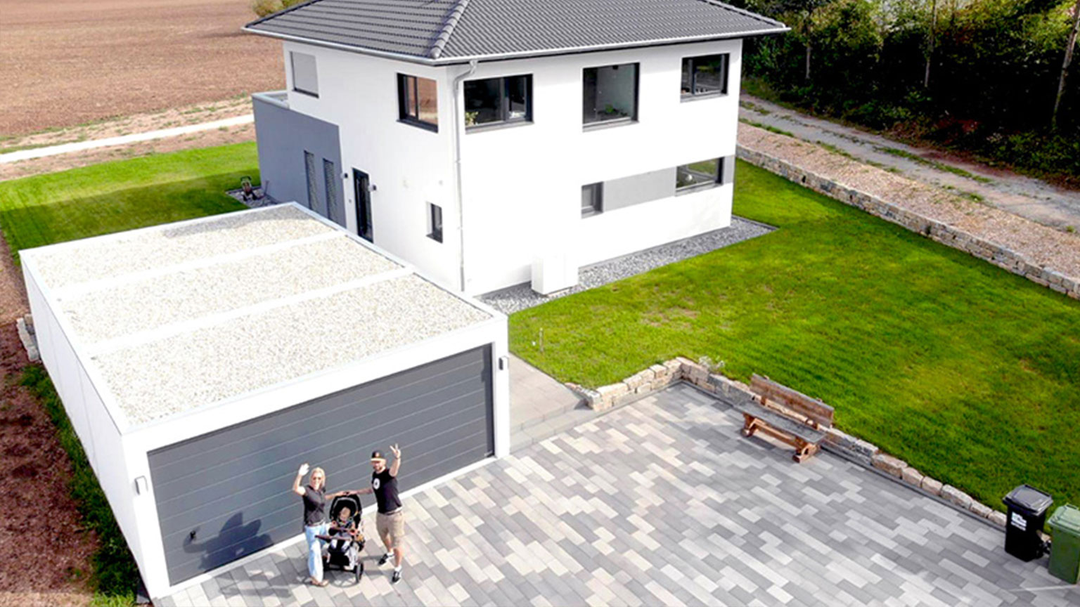 Luftaufnahme eines modernen Einfamilienhauses mit Doppelgarage und gepflasterter Einfahrt, eine Familie mit Kinderwagen winkt in die Kamera.