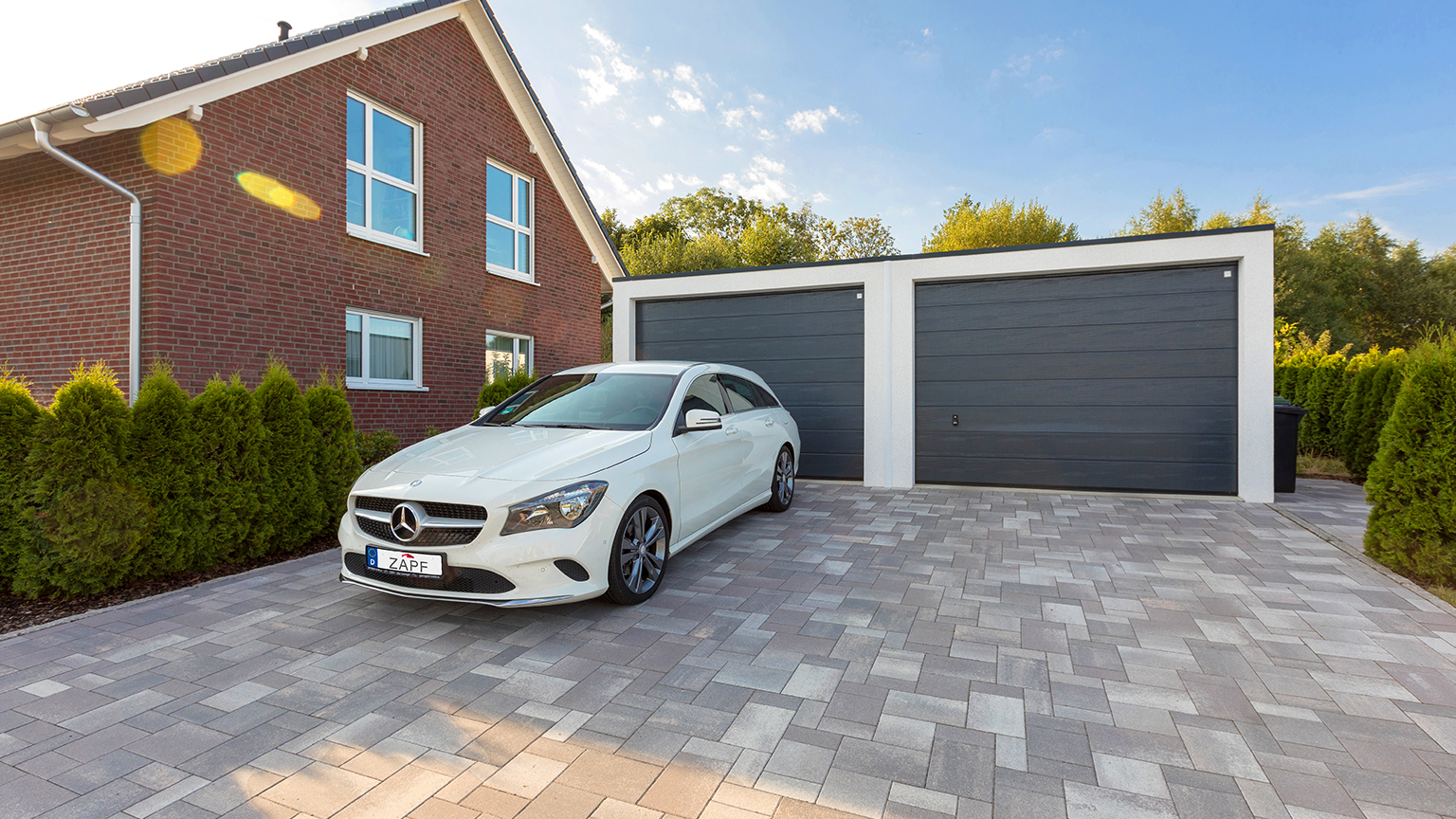 Weißer Mercedes vor moderner Doppelgarage mit dunklen Sektionaltoren auf gepflasterter Einfahrt.