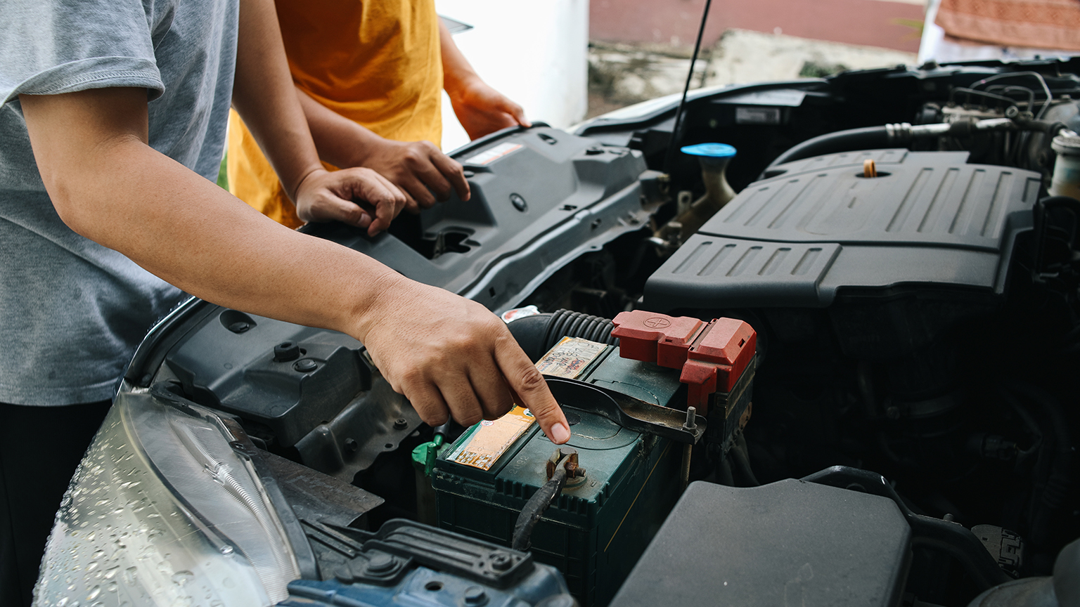 Zwei Personen prüfen gemeinsam eine Autobatterie unter geöffneter Motorhaube bei Tageslicht.