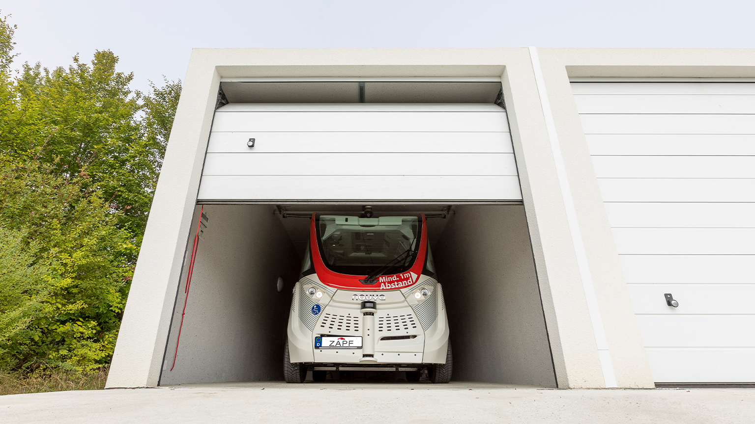 Selbstfahrender Minibus in moderner Fertiggarage mit leicht geöffnetem Sektionaltor und grüner Umgebung.