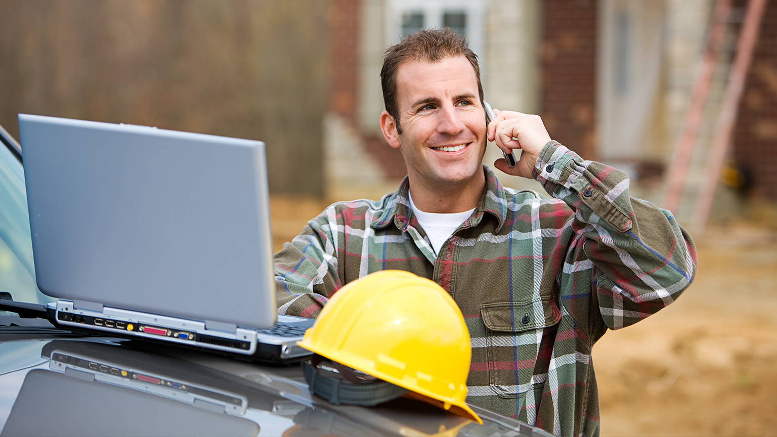 Bauleiter mit Laptop und Schutzhelm telefoniert vor Baustelle mit Rohbau im Hintergrund.