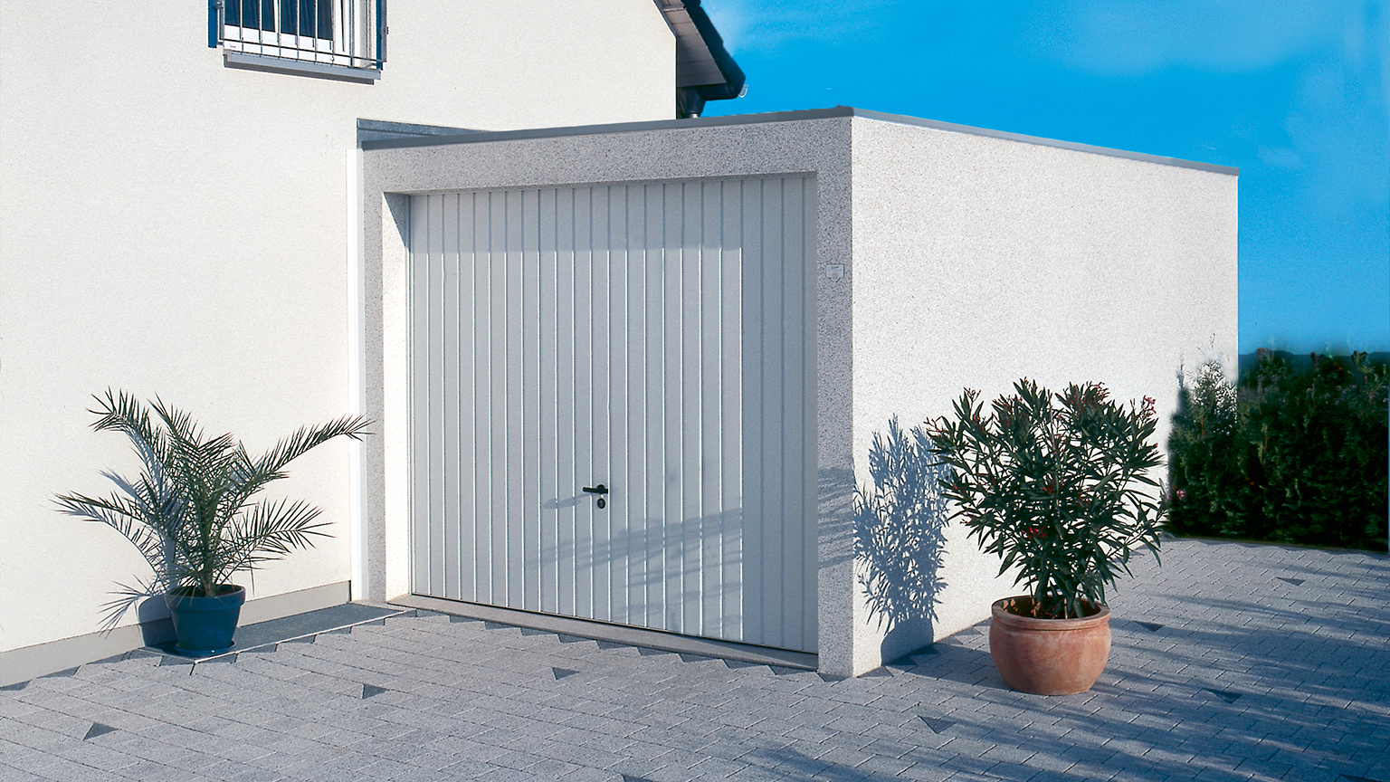 Einzelgarage mit weißem Schwingtor neben Wohnhaus, eingerahmt von Kübelpflanzen.