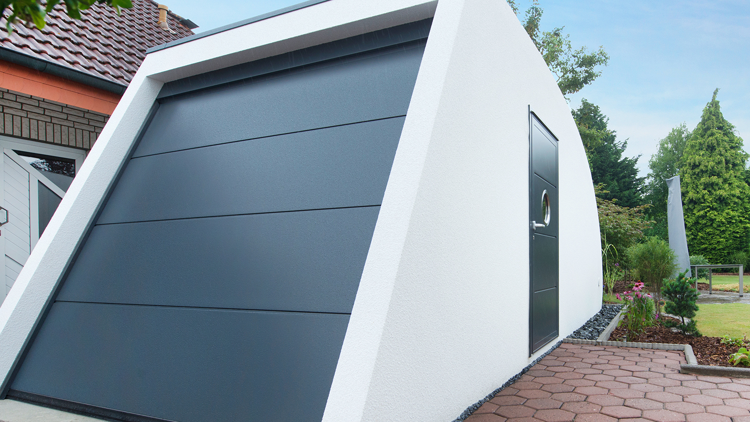 Moderne Trapezgarage mit schräger Seitenwand und dunklem Sektionaltor vor Wohnhaus.