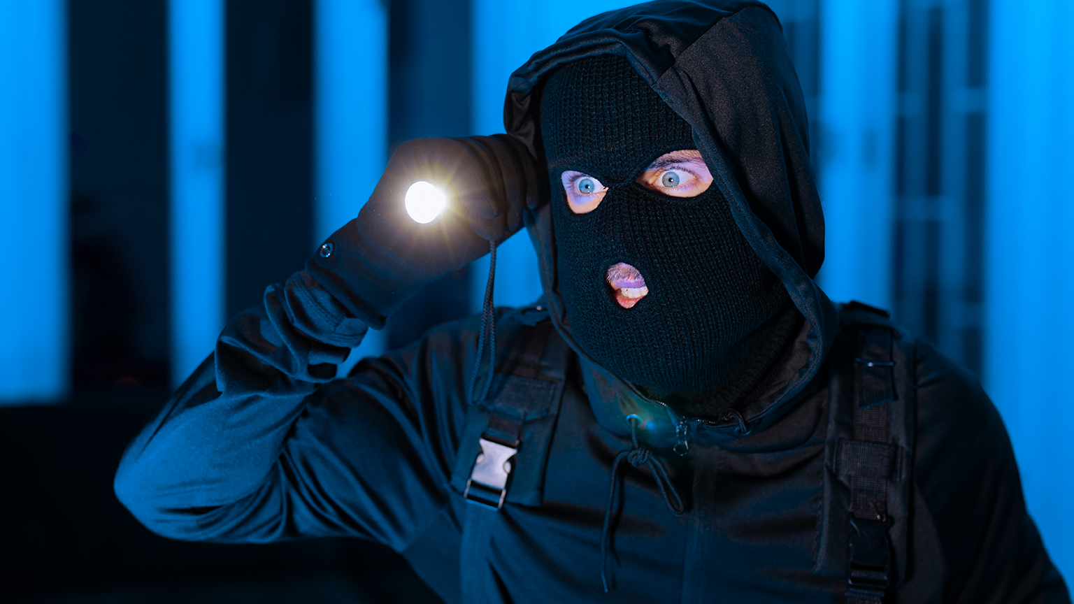 Maskierter Einbrecher mit Sturmhaube und Taschenlampe bei Nacht im blauen Licht.