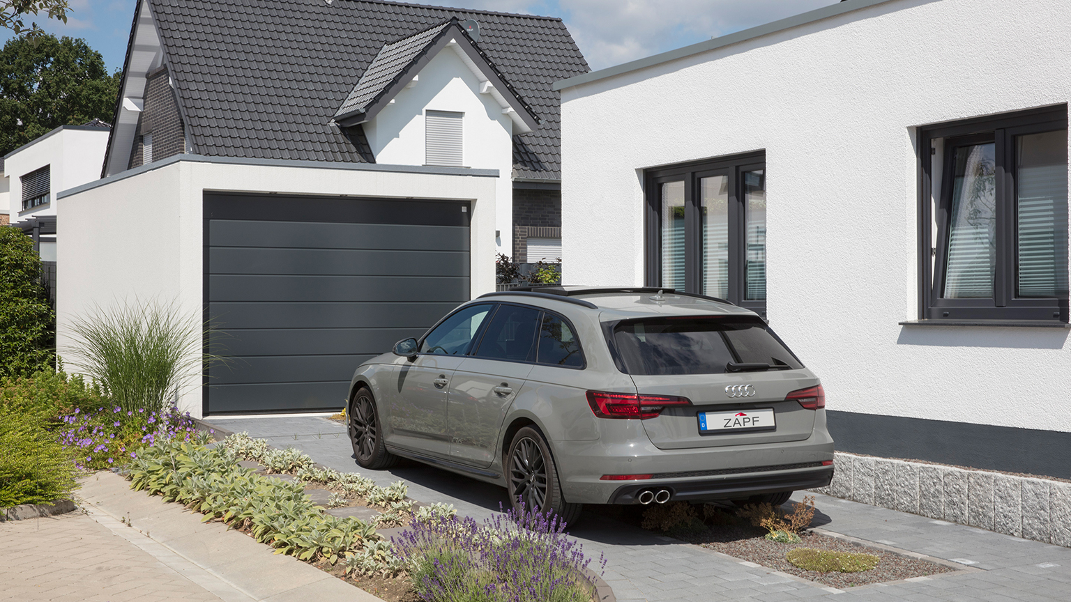 Ein Einfamilienhaus mit Garage, dunklem Garagentor und geparktem Auto in einer bepflanzten Einfahrt bei Sonnenschein.
