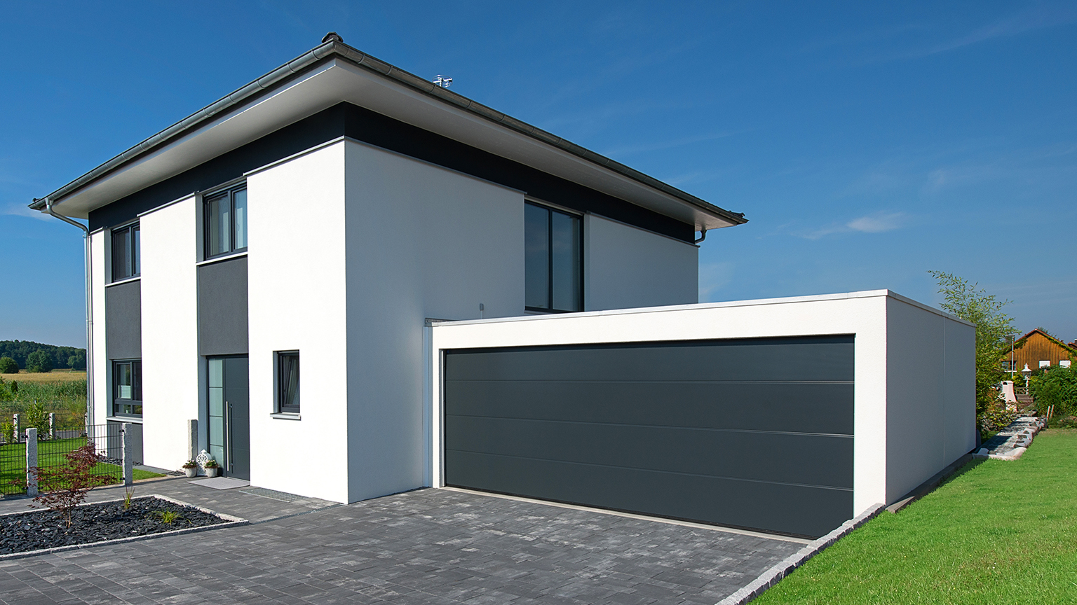 Moderne weiße Stadtvilla mit Flachdach-Doppelgarage in Anthrazit; die Garage ist seitlich ans Haus angebaut, mit breiter Einfahrt und gepflasterter Hofeinfahrt bei sonnigem Wetter.