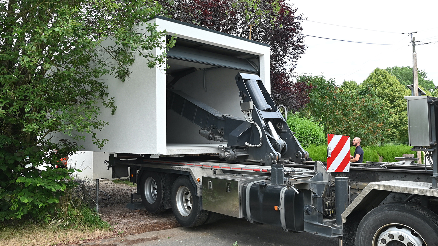 Anlieferung einer Fertiggarage per LKW mit hydraulischem Hebearm auf eine Baustelle im Grünen.