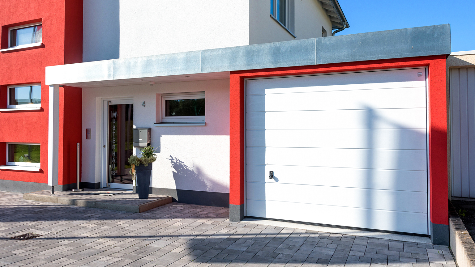 Eingangsbereich eines modernen Musterhauses mit integrierter weißer Garage und rotem Fassadenakzent.