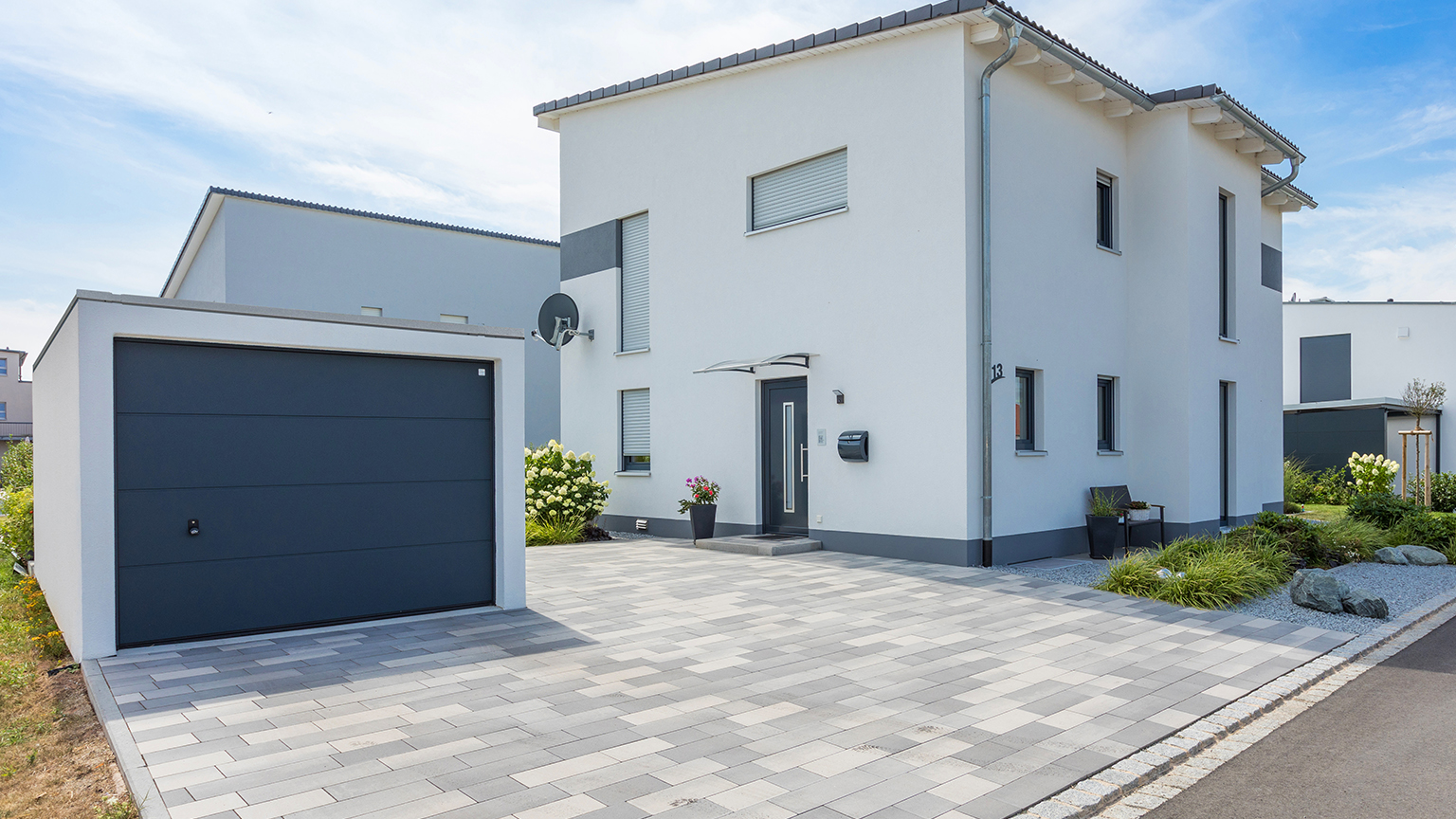 Neubau mit weißer Fassade, dunklen Fensterrahmen und Garage samt strukturierter Einfahrt und Vorgartenbegrünung.