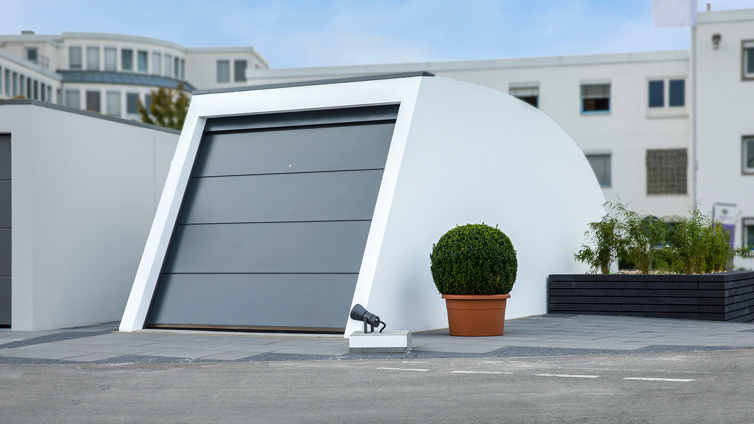Design-Garage mit futuristischer Form und grauem Sektionaltor vor moderner Büroarchitektur.