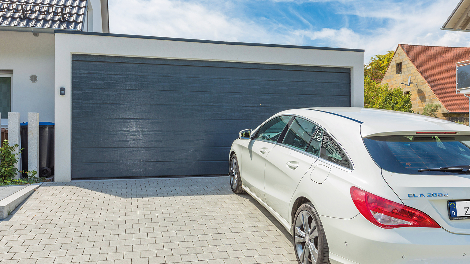 Weißes Auto vor moderner Garage mit anthrazitfarbenem Sektionaltor auf gepflasterter Einfahrt.