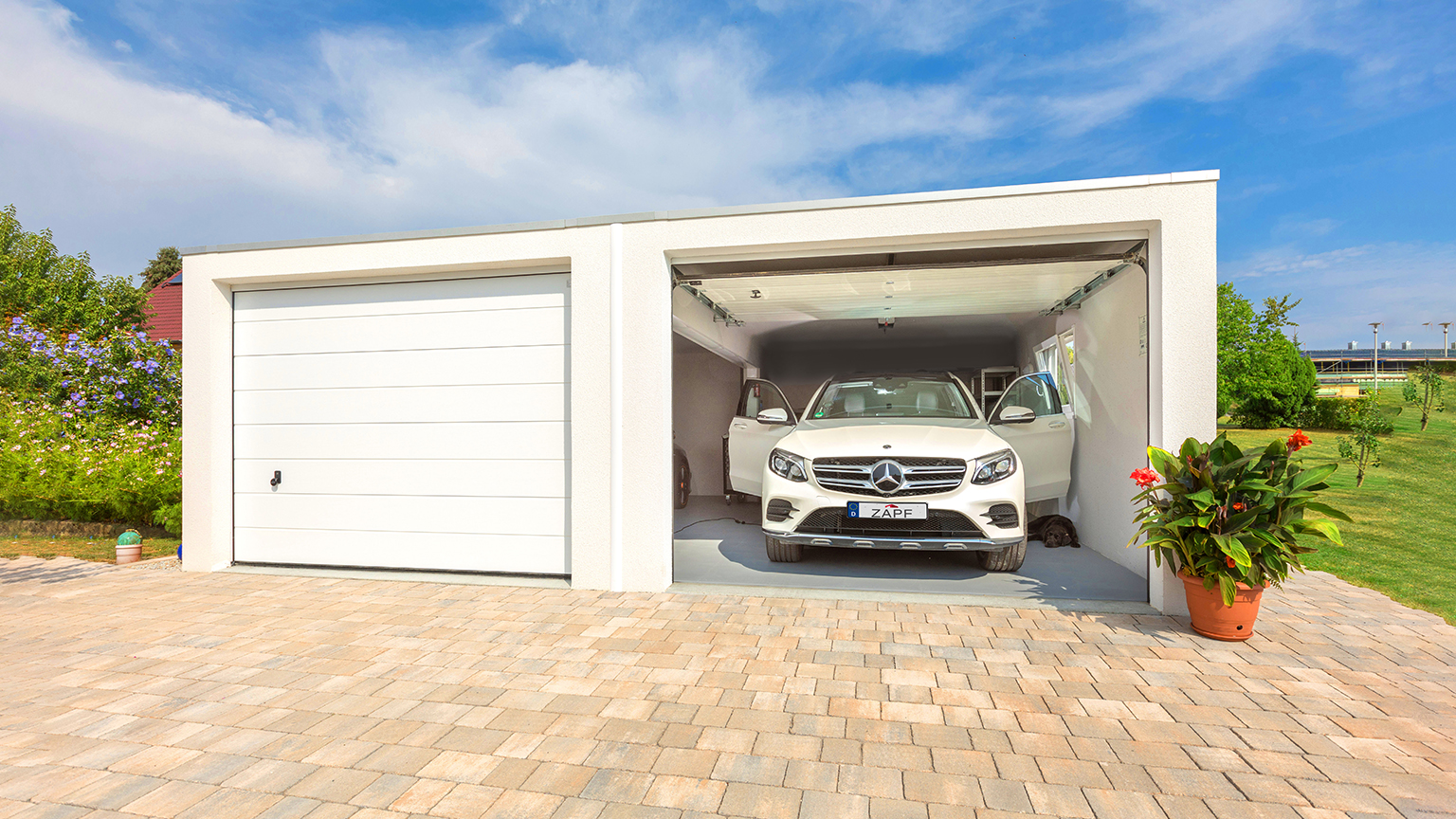 Weiße Doppelgarage mit geöffnetem Tor und geparktem Mercedes-SUV auf gepflasterter Einfahrt im Grünen.