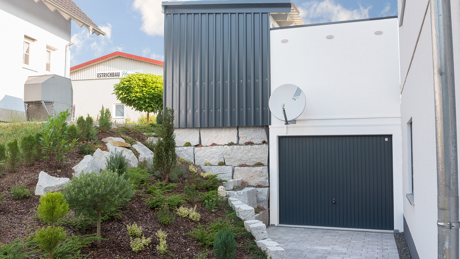 Hanglage mit moderner Fertiggarage und Satellitenschüssel, umgeben von bepflanztem Garten und Natursteinmauer.