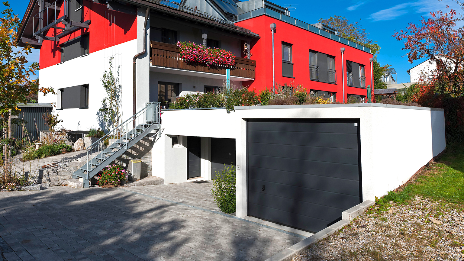 Moderne weiße Doppelgarage mit schwarzem Sektionaltor vor stilvollem Wohnhaus mit roter Fassade und Gartenterrasse.