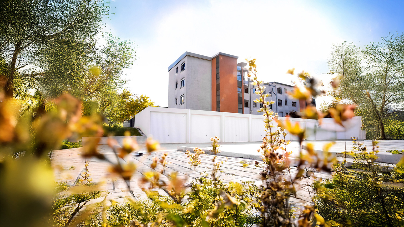Weiße Reihengaragen vor mehrstöckigem Wohnhaus in begrünter Wohnanlage bei Sonnenschein.