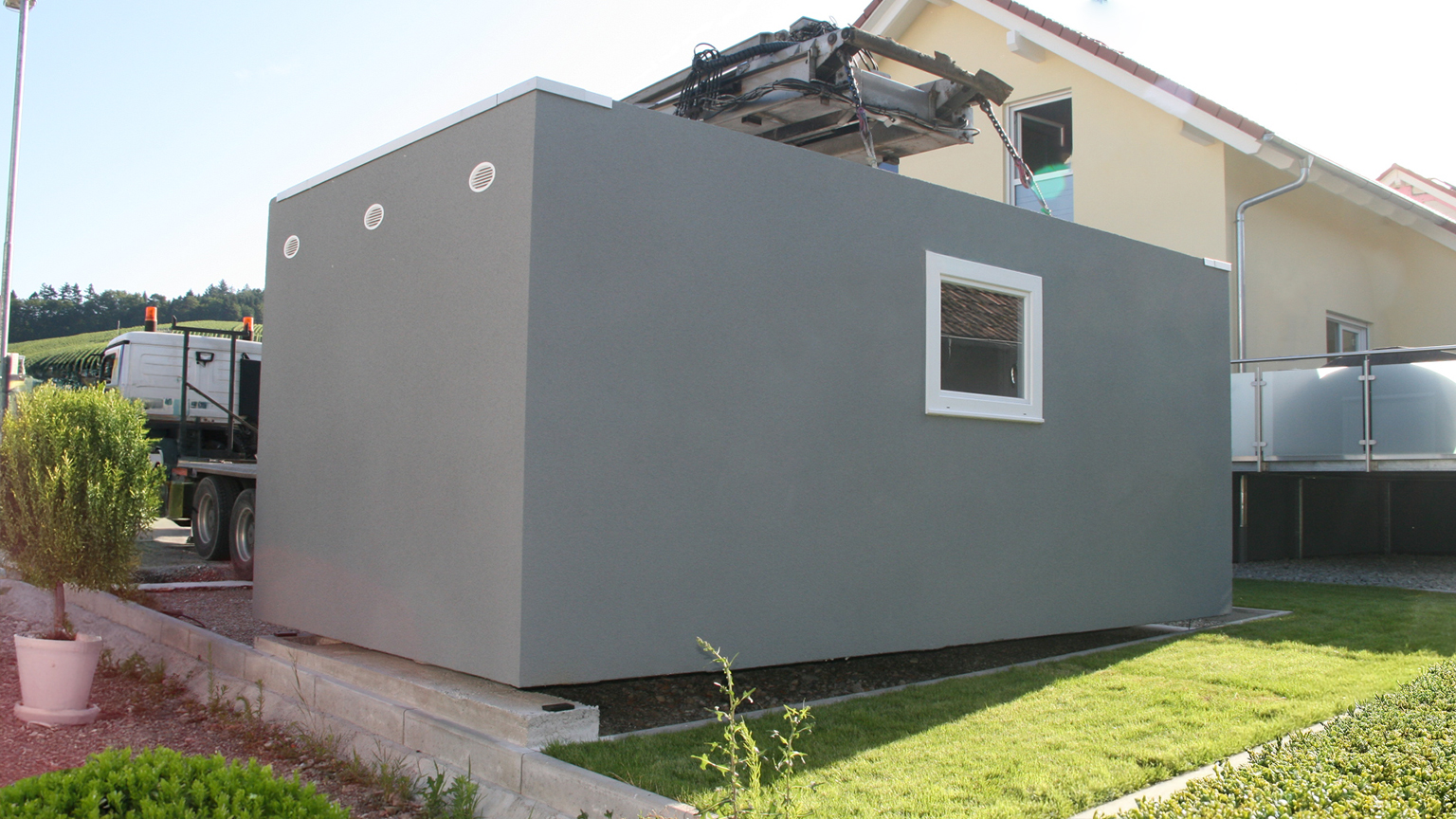 Fertiggarage in moderner grauer Farbe wird per Kran an ihren endgültigen Standort neben einem Wohnhaus gesetzt