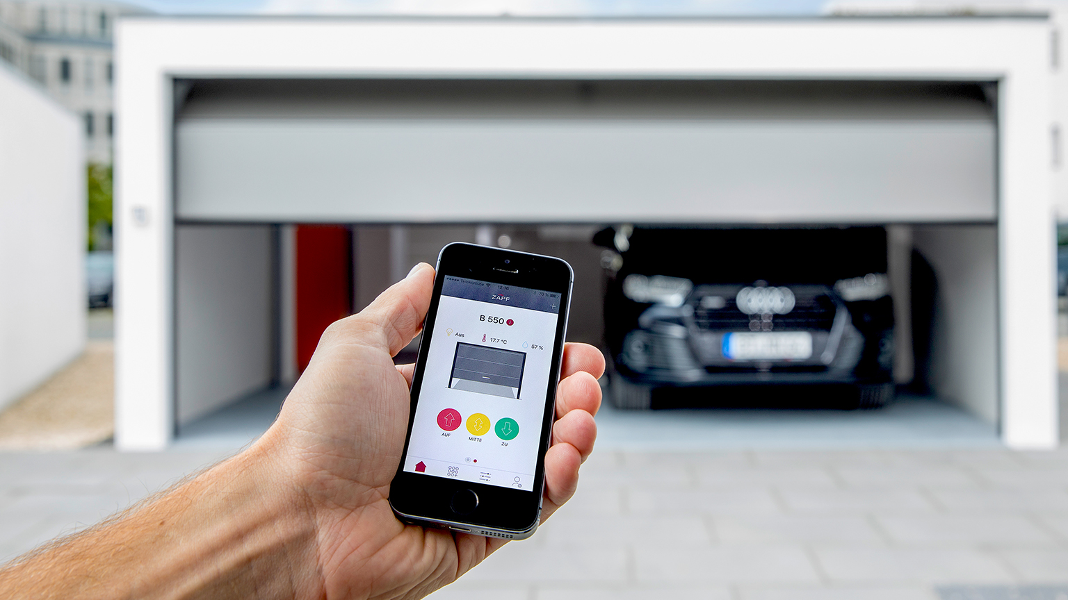 Smartphone steuert Garagentor per App, im Hintergrund moderne Fertiggarage mit schwarzem Audi.