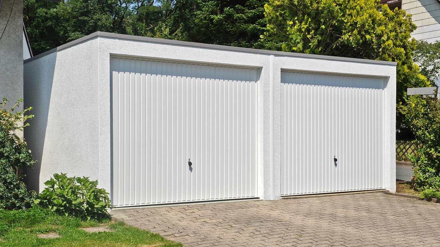Doppelgarage mit weißem Schwingtor und Putzfassade, eingebettet in grüne Gartenumgebung.