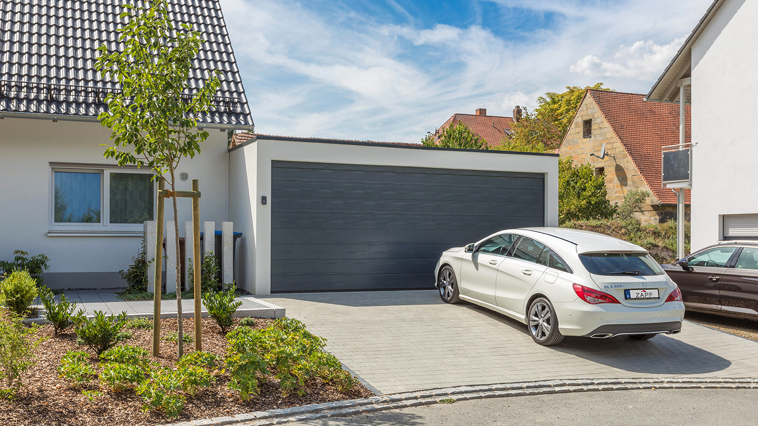 Moderne Doppelgarage mit dunklem Sektionaltor und weißem Mercedes auf gepflasterter Einfahrt.