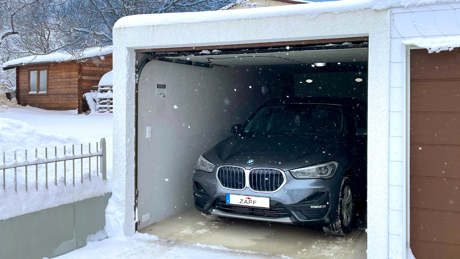 BMW mit ZAPF-Kennzeichen steht in geöffneter Garage bei Schneefall und winterlicher Kulisse mit Holzhaus.