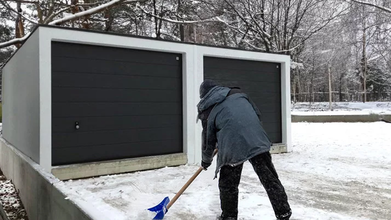 Winterliche Szene mit schneebedecktem Boden und einer Person, die vor zwei geschlossenen Garagentoren Schnee schaufelt.