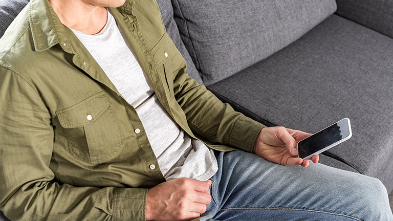 Mann sitzt entspannt auf einem Sofa und hält ein Smartphone in der Hand.
