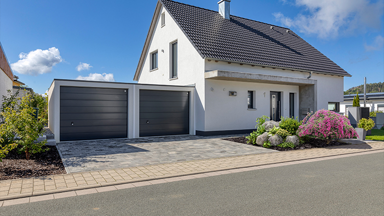Moderne Doppelgarage von ZAPF neben einem Einfamilienhaus mit gepflegtem Vorgarten und gepflasterter Einfahrt bei sonnigem Wetter.