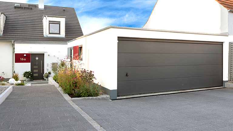 Garage double moderne blanc avec porte sectionnelle anthracite sur un terrain bien entretenu devant une maison individuelle.