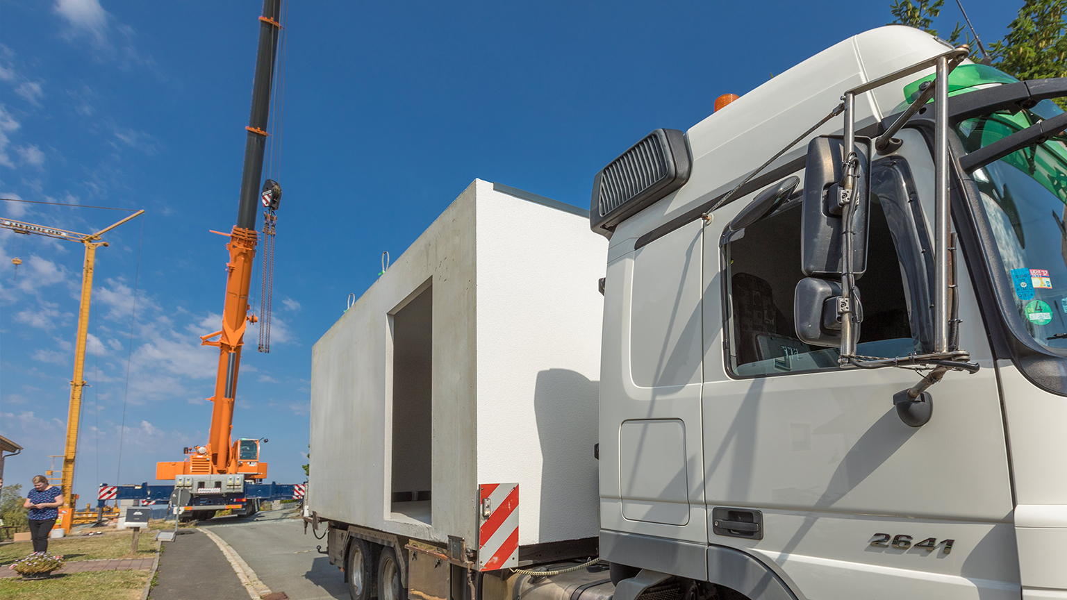Kran hebt Fertiggarage von LKW bei Anlieferung auf Baustelle unter blauem Himmel.