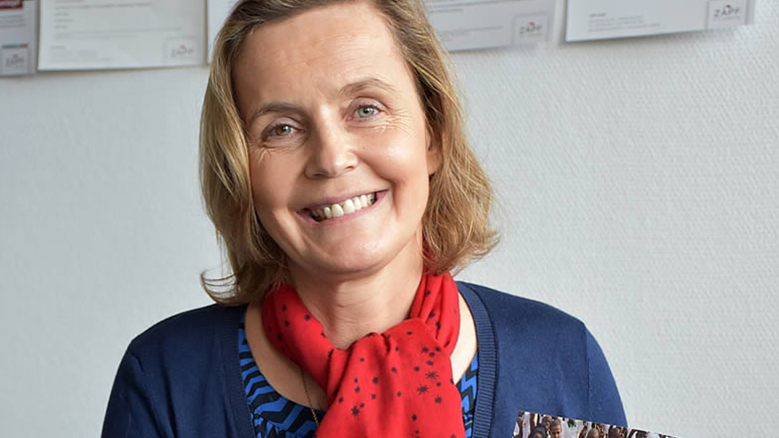Freundlich lächelnde Frau mit rotem Schal vor weißer Wand im Büro mit ZAPF Dokumenten im Hintergrund.