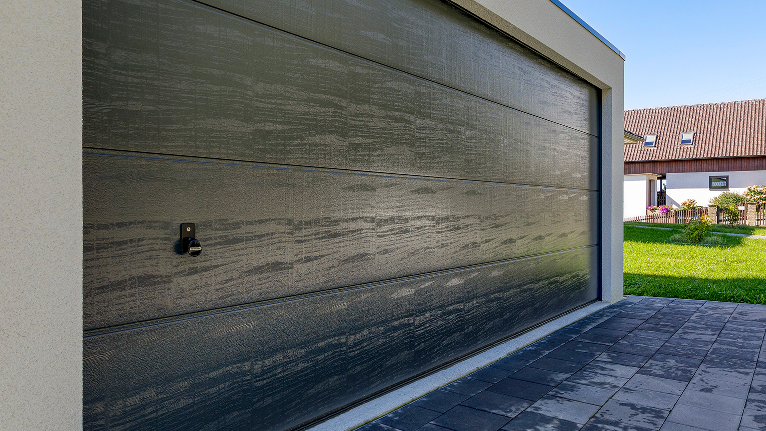 Moderne Fertiggarage mit dunklem Sektionaltor in Holzoptik vor einem Wohnhaus im Grünen.