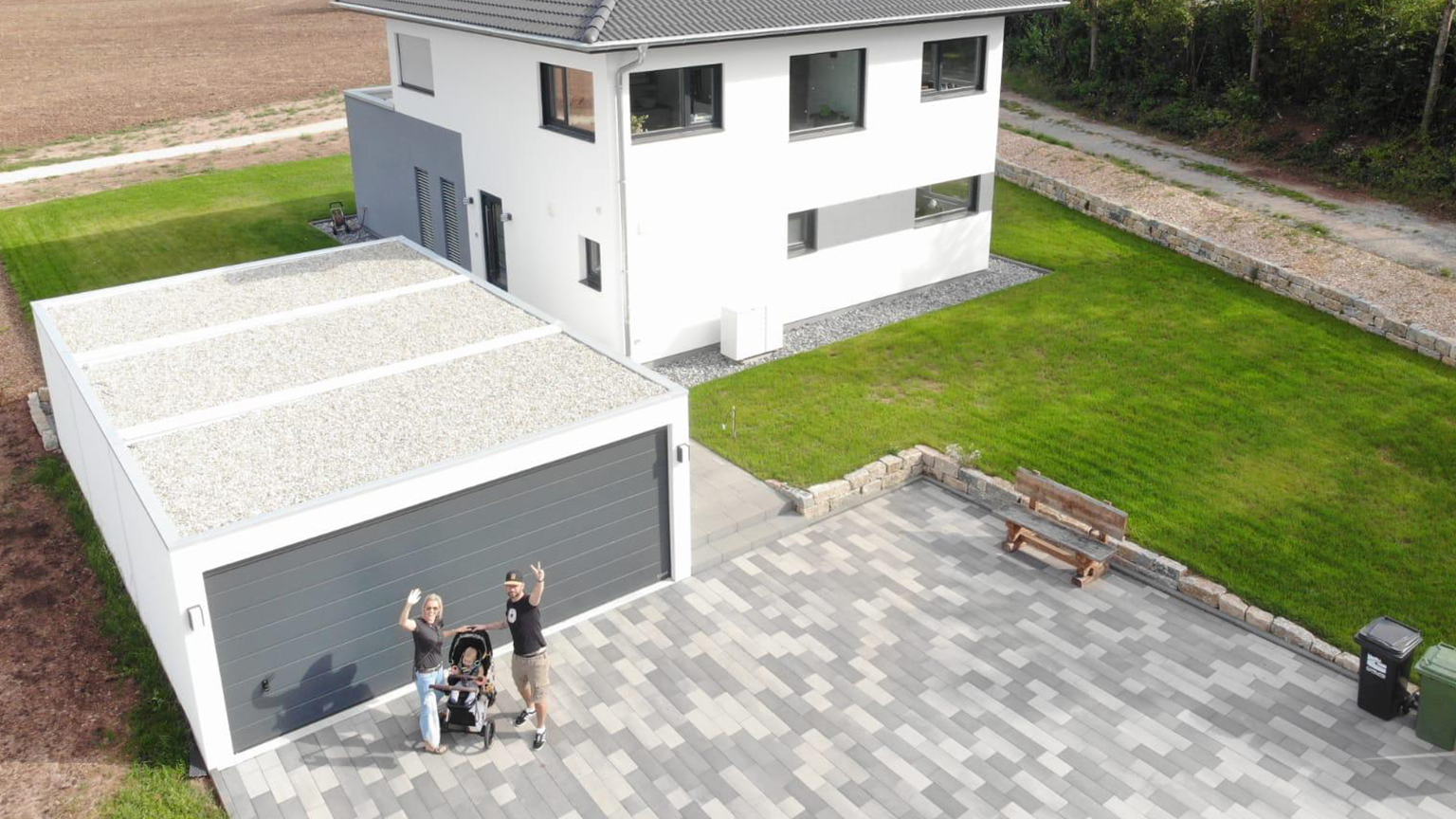 Familie mit Kinderwagen vor moderner ZAPF Großraumgarage und weißem Einfamilienhaus aus der Vogelperspektive.