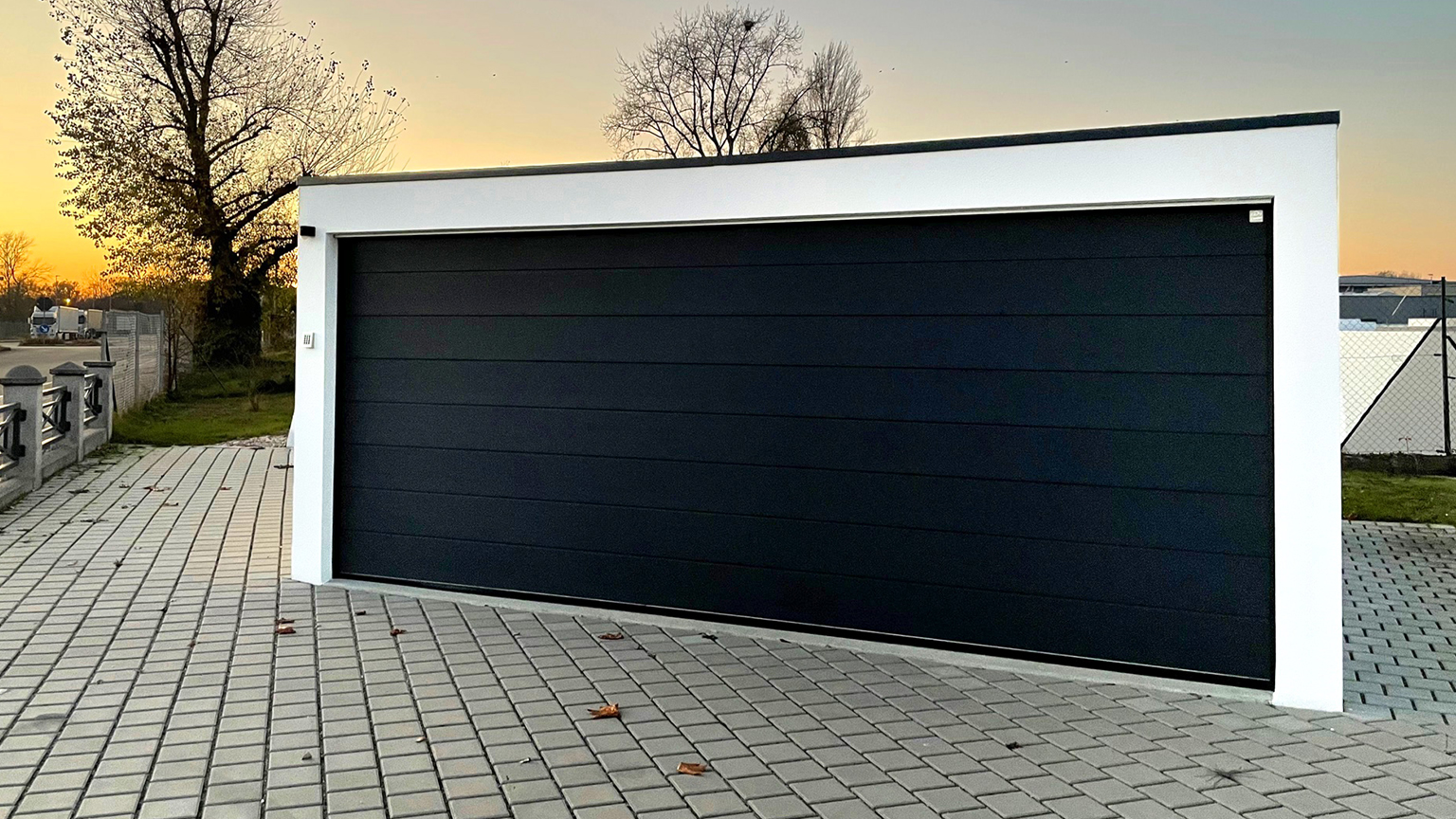 Moderne Großraumgarage mit dunklem Sektionaltor im Abendlicht auf gepflasterter Fläche.