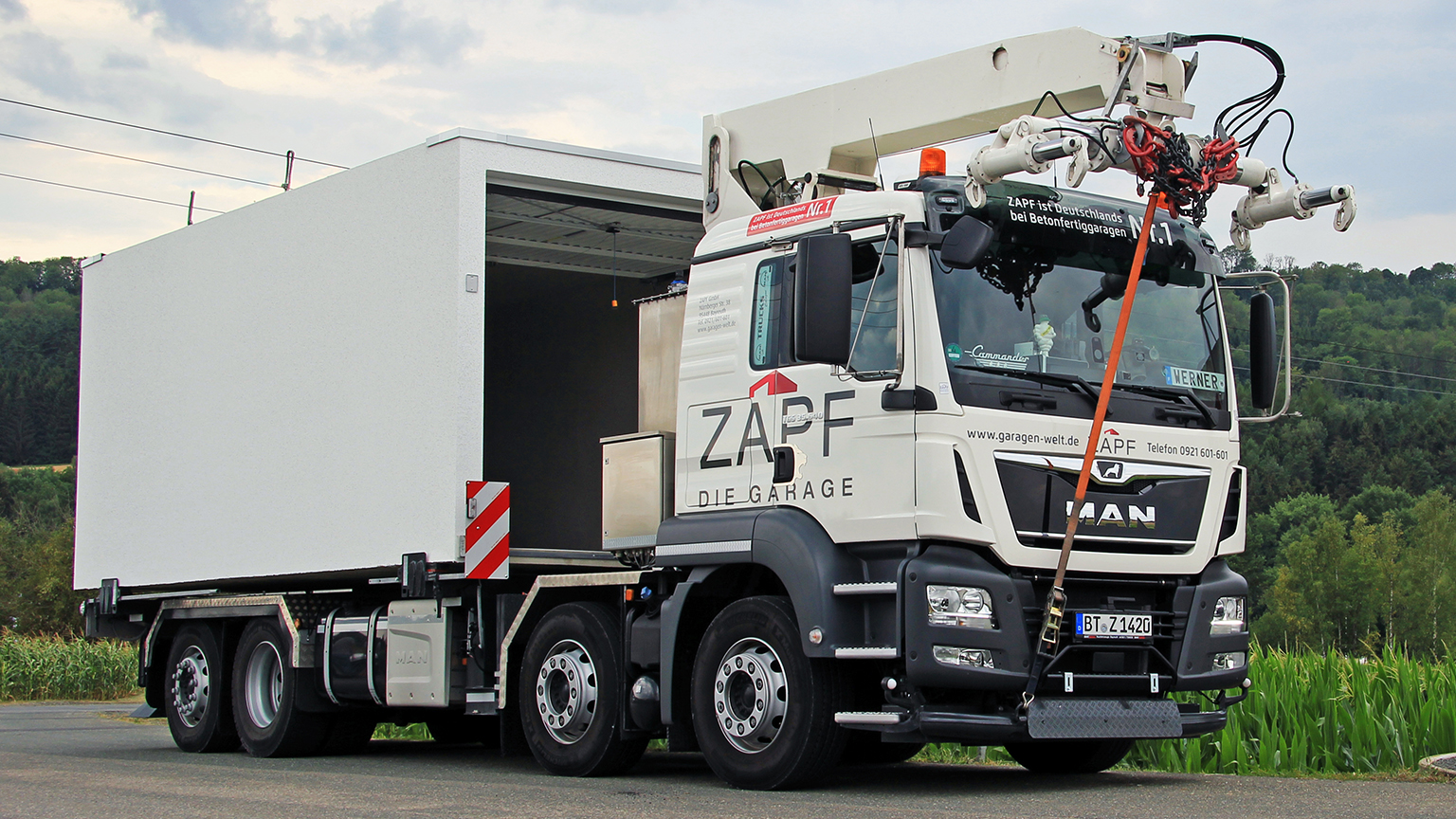 ZAPF LKW mit Spezialkran transportiert eine Fertiggarage vor grünem Landschaftshintergrund.