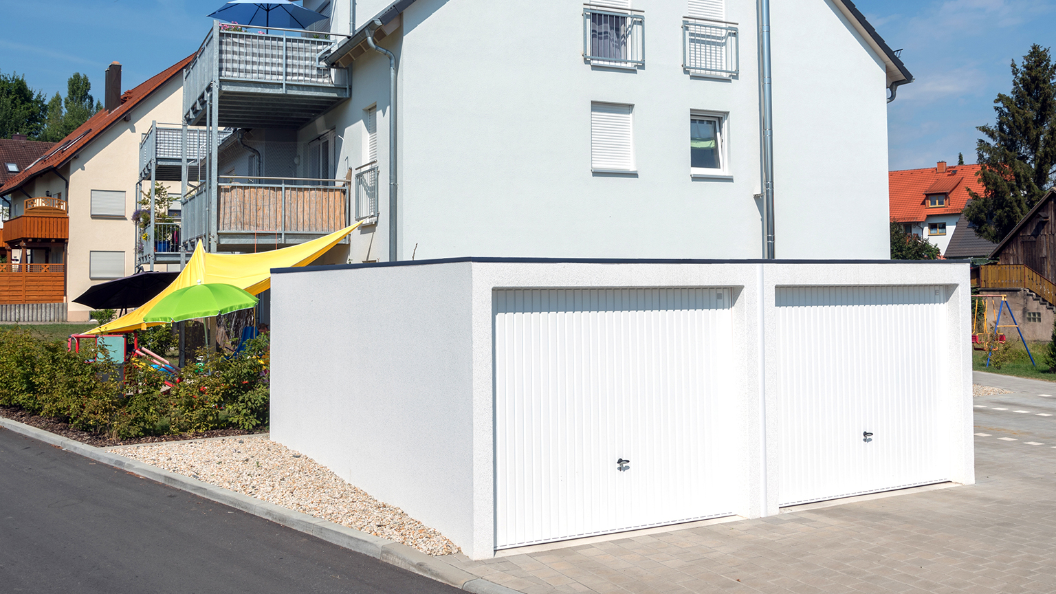 Weiße Doppelgarage neben einem Mehrfamilienhaus mit Spielplatz und bunten Sonnensegeln.