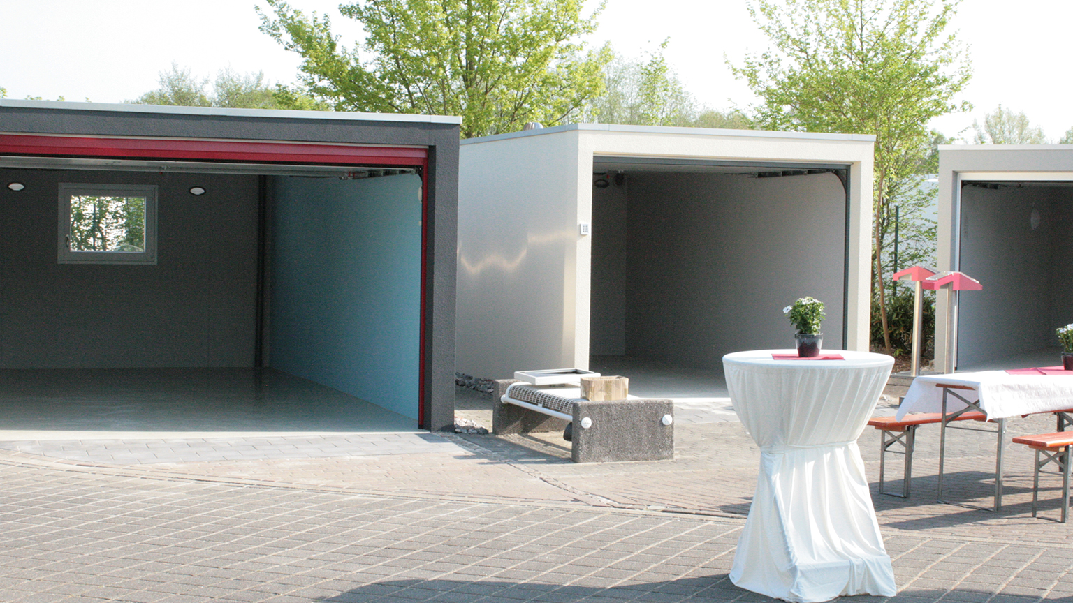 Offene Muster-Garagen in verschiedenen Farben bei einer ZAPF Ausstellung im Freien, dekoriert mit Stehtischen und Sitzbänken.