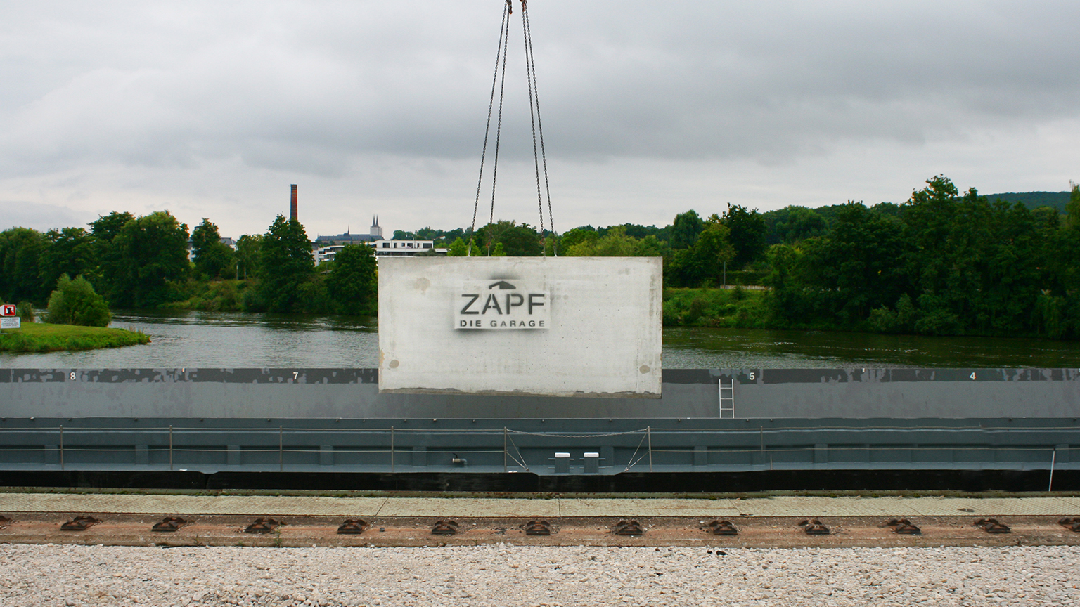 Betongarage mit ZAPF Logo wird per Kran auf ein Binnenschiff am Flussufer verladen.
