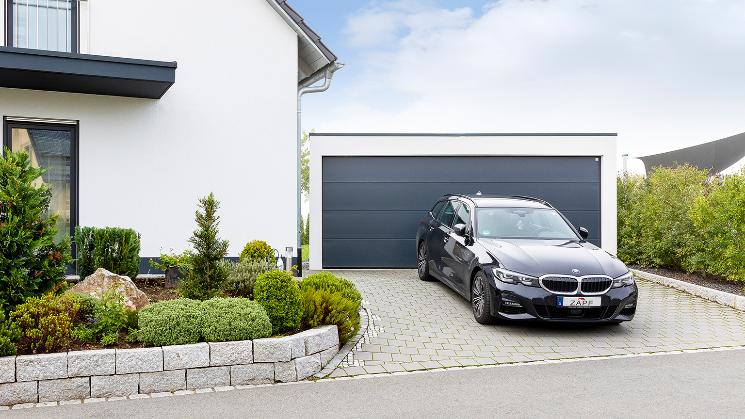 Schwarzer BMW steht vor moderner ZAPF Fertiggarage mit dunklem Sektionaltor und gepflegtem Vorgarten.