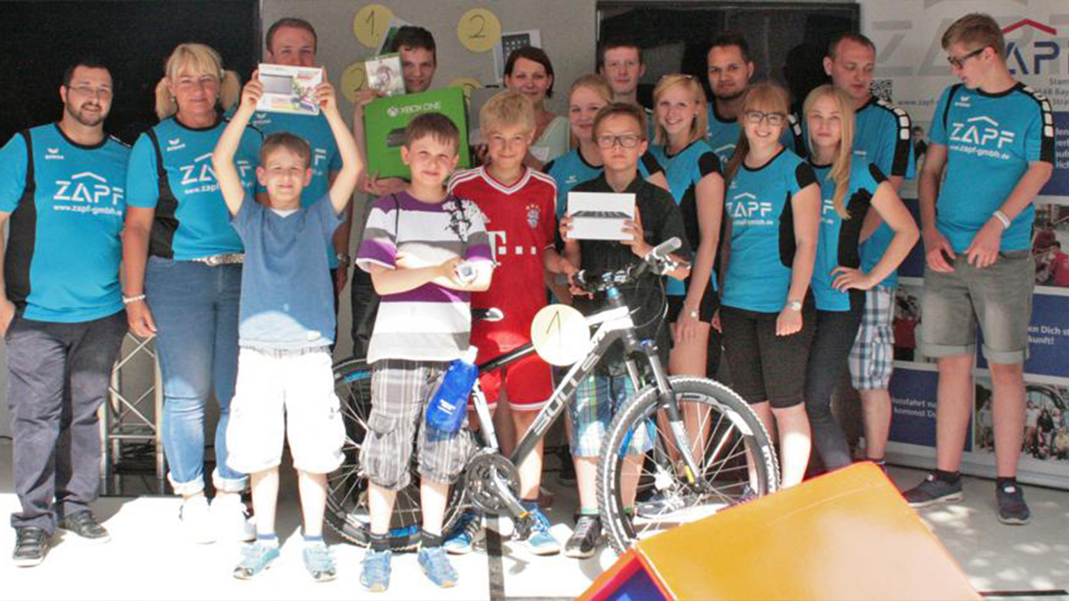 Gruppenfoto von ZAPF- itarbeitern und Kindern bei einer Preisverleihung mit Sachgewinnen wie Fahrrad, Konsole und Tablet.