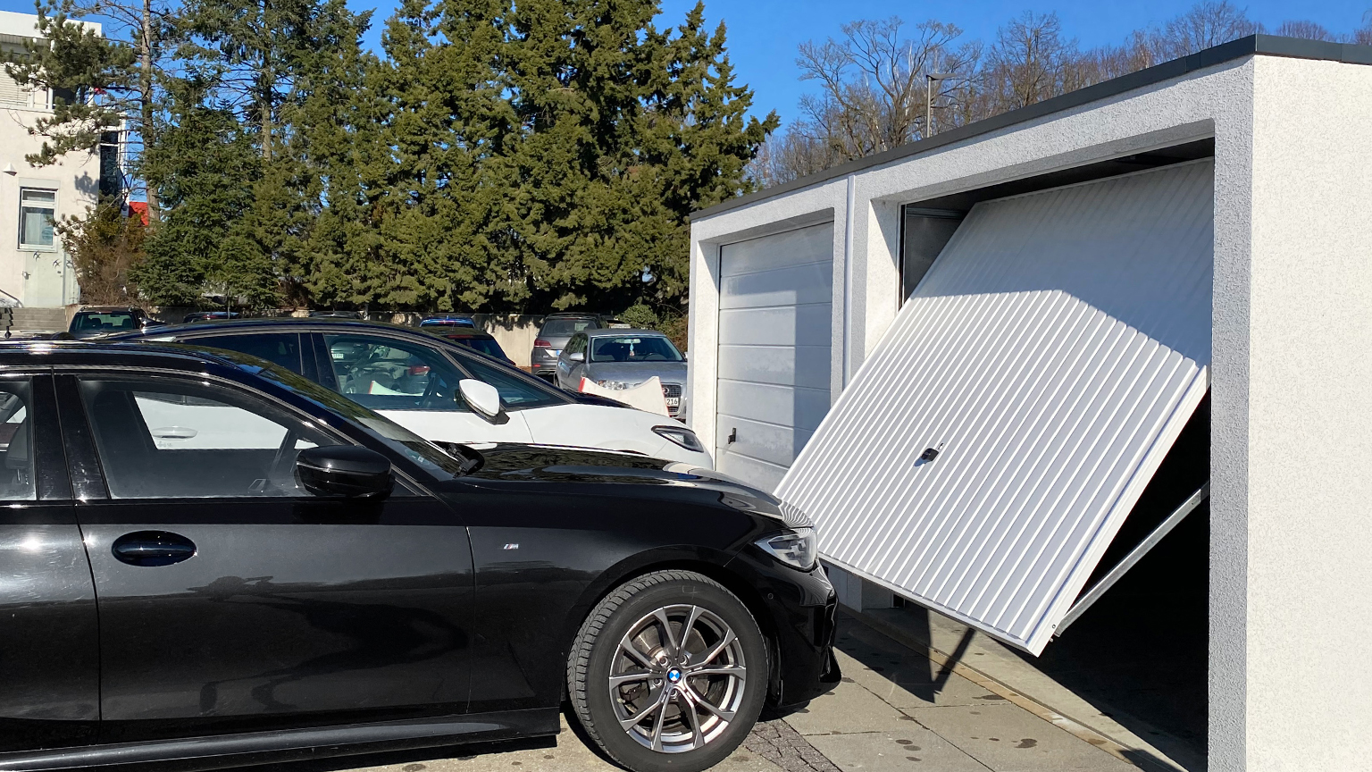 Schwarzer BMW vor weißer Fertiggarage mit geöffnetem Schwingtor bei Sonnenschein.