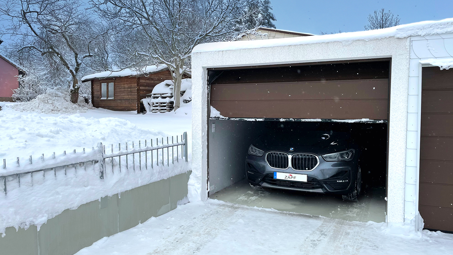 Schneebedeckte Fertiggarage mit halb geöffnetem Tor und geparktem BMW X1 im Winter.